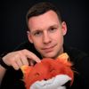 Portrait of a person in a black shirt holding an orange fox plush toy, looking directly at the camera against a dark background.