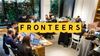 Busy workshop scene with around a dozen people gathered in a co-working space. Participants sit and stand around wooden tables covered with laptops, notebooks, coffee cups, and cables while collaborating on projects. Large windows along one wall look out onto dense green foliage, creating an open and welcoming atmosphere. A prominent yellow banner displaying the word FRONTEERS spans the middle of the image.