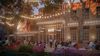 Evening view of a lively courtyard outside a modern glass-fronted venue. String lights hang between large trees while groups of people sit and socialize at outdoor tables surrounded by colourful plants and flower beds.