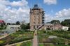 A former water tower rises above landscaped gardens and greenhouses. Vegetable beds, pathways, and historic brick buildings fill the foreground under a bright sky with scattered clouds.