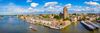 Panoramic aerial view of Dordrecht on a sunny day. The river curves through the city, with boats on the water, historic buildings along the waterfront, and the prominent tower of the Grote Kerk rising above the skyline beneath scattered clouds.